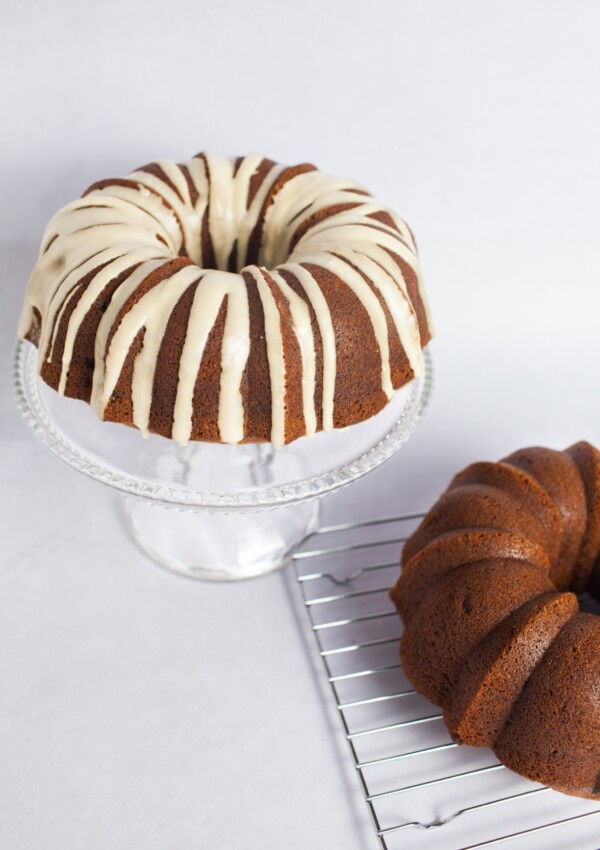 Glazed Banana Rum Raisin Cake on a raised cake plate with a plain cake on a wire rack half visible in the foreground