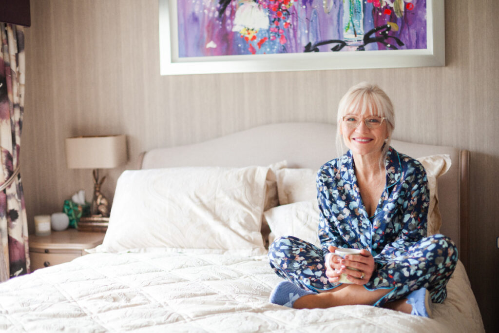 April Harris wearing pyjamas sitting cross-legged on a bed holding a cup of coffee
