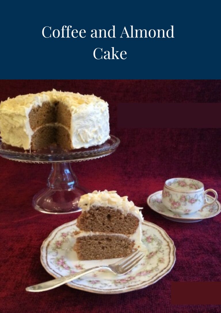 A coffee and almond cake on a glass cake stand with a slice of cake in the foreground on a china plate. There's a tea cup in the background.