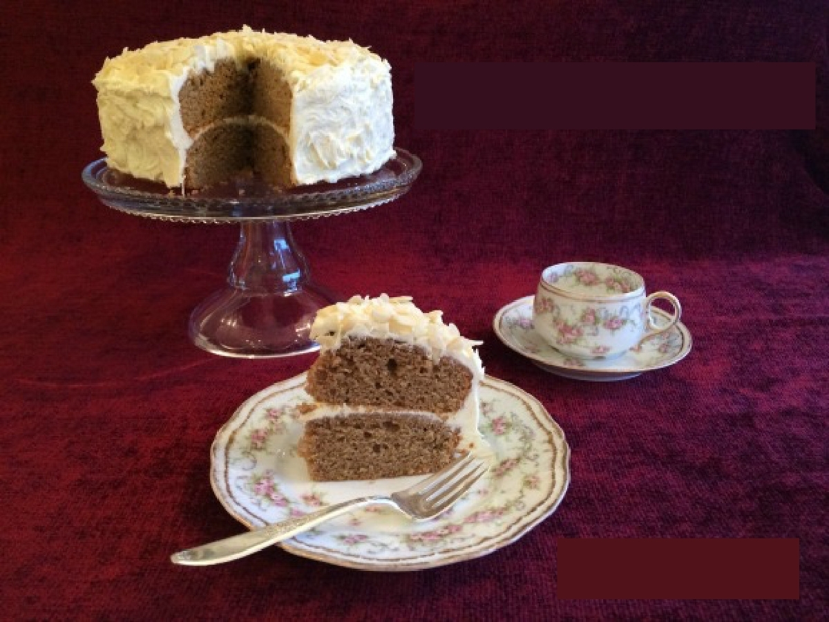 A coffee and almond cake on a glass cake stand with a slice of cake in the foreground on a china plate. There's a tea cup in the background.