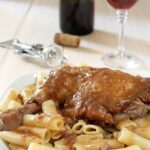 Pastitsada served over pasta with a glass of red wine in the background