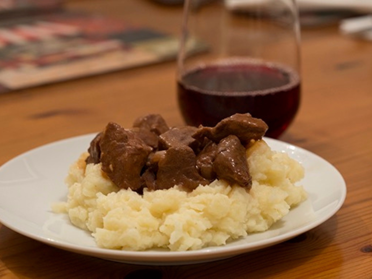 Poor Man's Steak Diane served over mashed potatoes on a white plate with a glass of red wine in the background