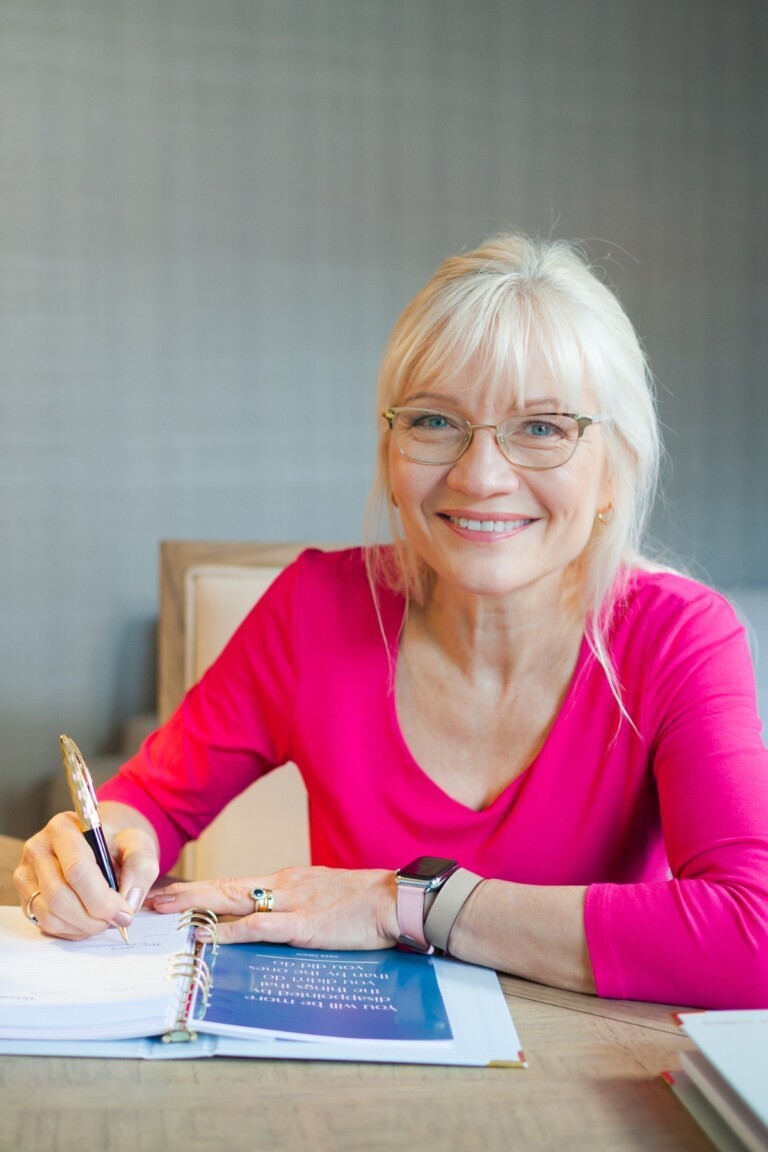 April Harris wearing a bright pink t-shirt, sitting and a desk and writing down her new years resolutions