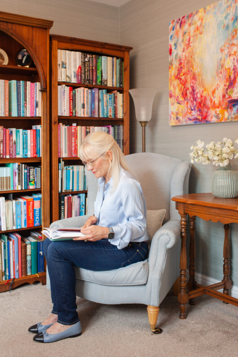 April J Harris sitting on a chair and reading a book in her library at home