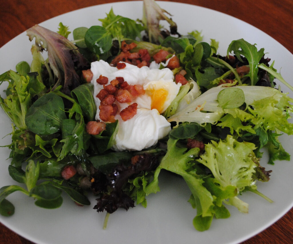 Salade Frisée with crispy bacon lardons, tangy greens and a poached egg served on a plate