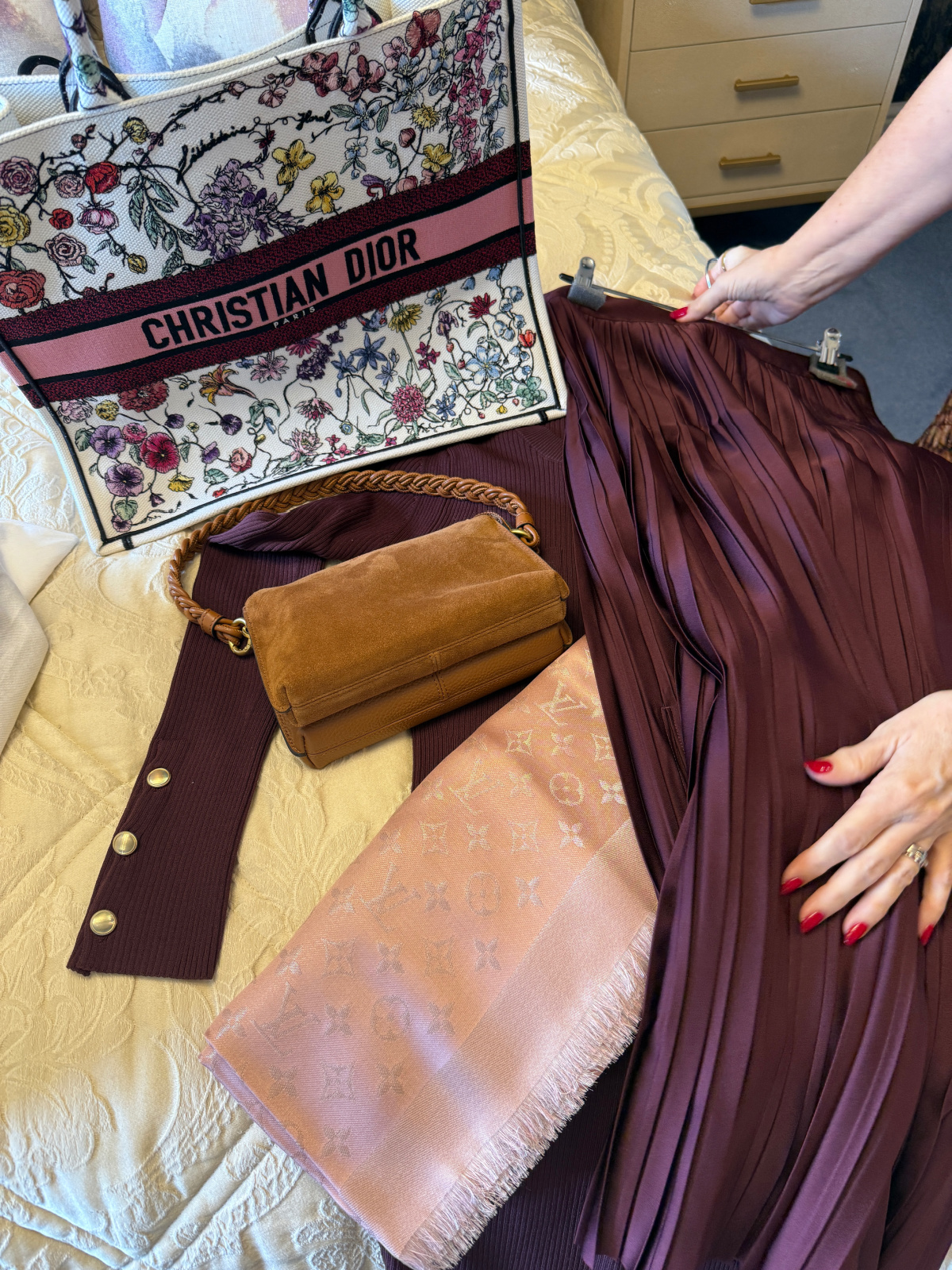 A flat lay of a burgundy dress and pleated skirt with a pink scarf, suede handbag and flowered Dior book bag