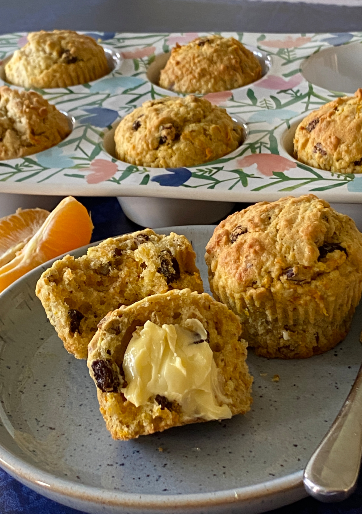Oat Bran Muffins with Carrot and Orange in a muffin tin and a plate holding a split muffin spread with butter in front.T