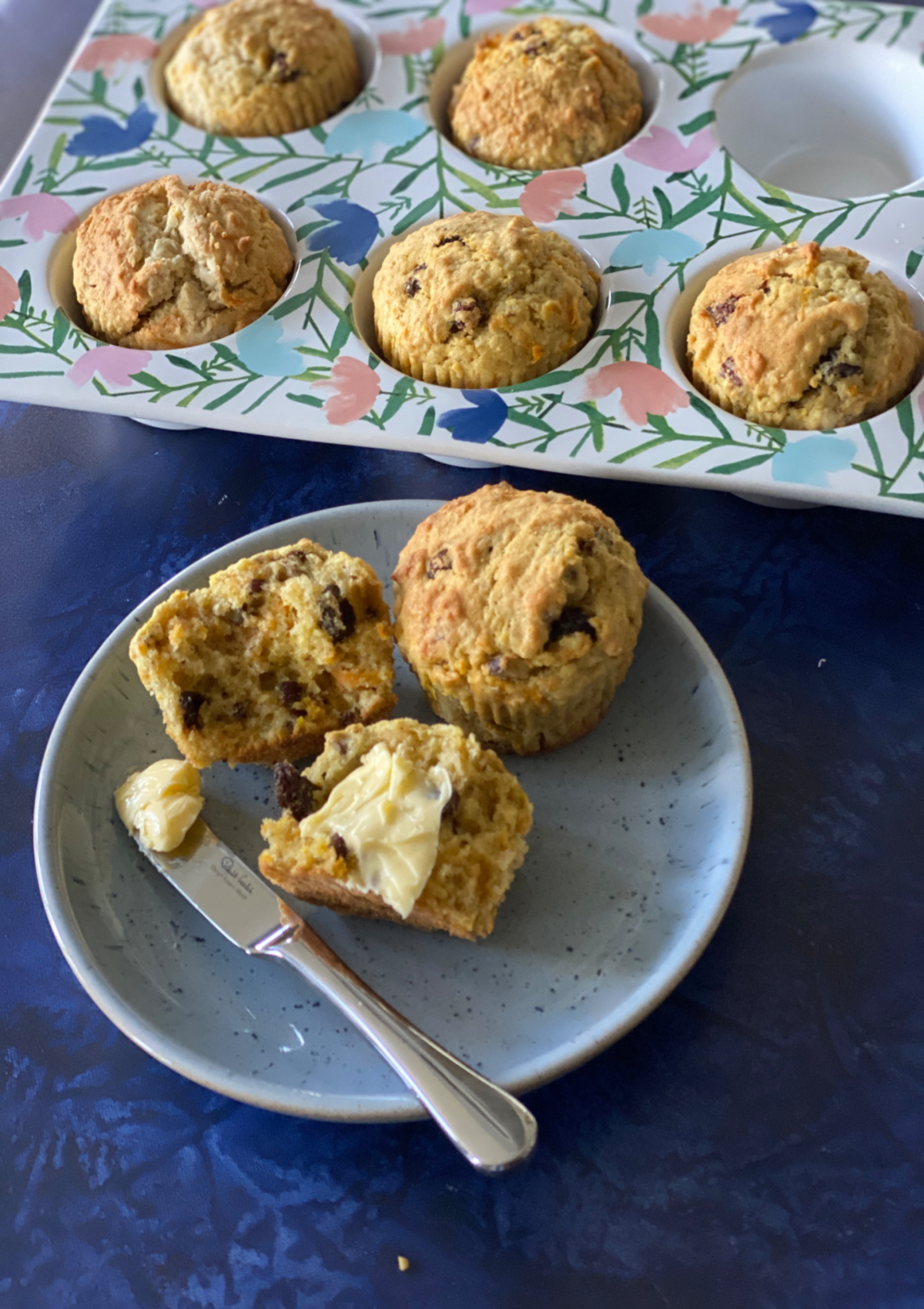 Oat Bran Muffins with Carrot and Orange in a muffin tin and a plate holding a split muffin spread with butter in front