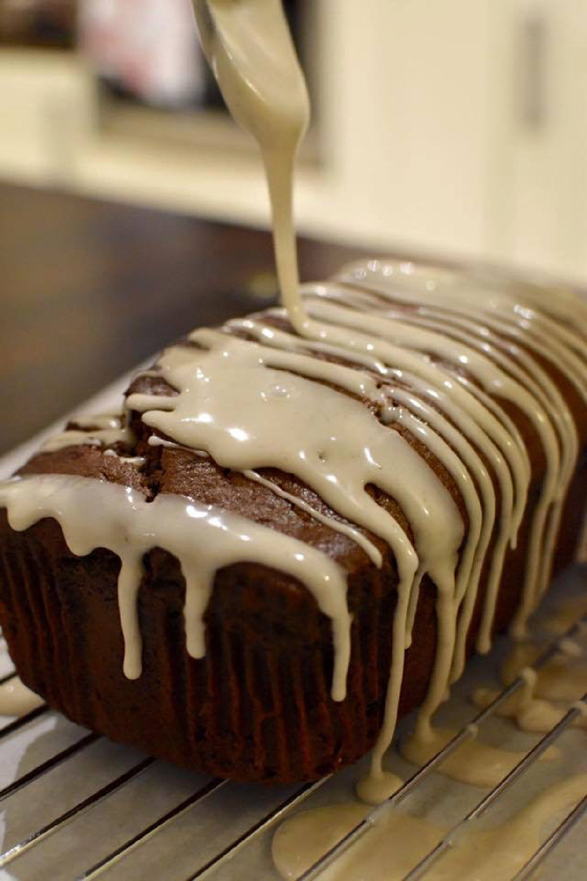 Banana Gingerbread Loaf Cake sitting on a wire rack as glaze is poured over top from a spoon