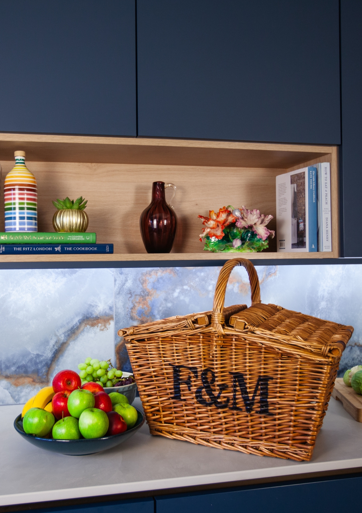 A kitchen counter with shelf above. There's a Fortnum and Mason basket and a bowl of fruit on the counter