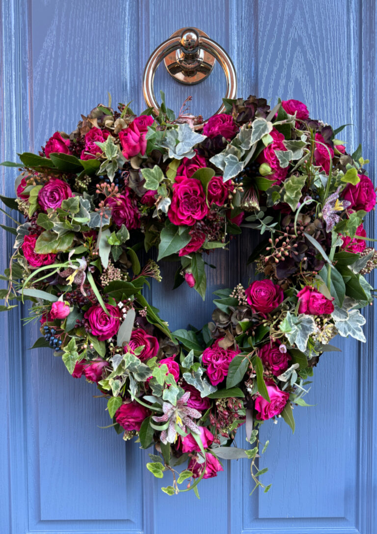 A heart shaped Valentine wreath made of roses and other live foliage hung on a blue front door