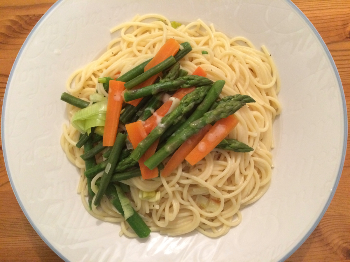 Spaghetti Primavera al Limone served on a white plate with a blue border. The spaghetti is topped with carrots, asparagus, leeks and other spring vegetables.