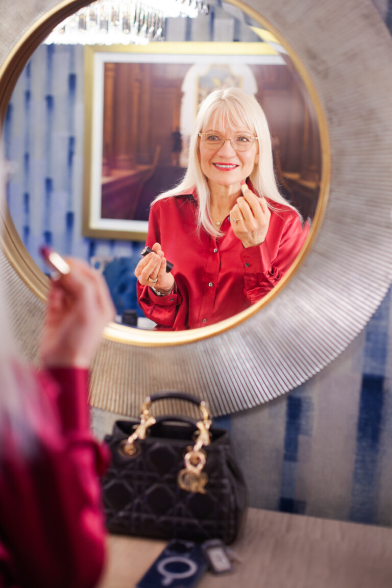 April Harris wearing a red blouse, looking into a mirror and applying lipstick