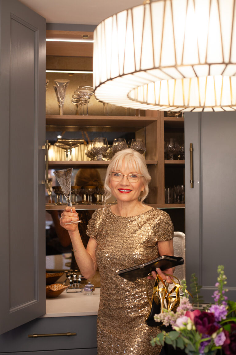 April Harris wearing a gold evening gown, taking a crystal champagne glass from her bespoke kitchen bar unit