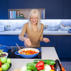 A photograph of April J Harris stirring sliced red peppers in a pan. There are fruit and vegetables arranged all around her on a kitchen island. There are blue cabinets behind her.