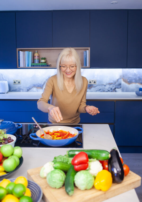 A photograph of April J Harris stirring sliced red peppers in a pan. There are fruit and vegetables arranged all around her on a kitchen island. There are blue cabinets behind her.