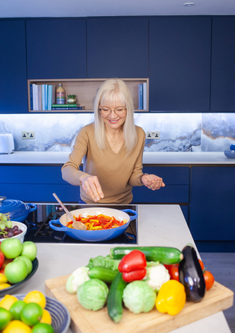 A photograph of April J Harris stirring sliced red peppers in a pan. There are fruit and vegetables arranged all around her on a kitchen island. There are blue cabinets behind her.