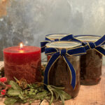 Three jars of suet-free mincemeat tied with blue and gold ribbon sitting beside a lit red candle encircled by Christmas foliage