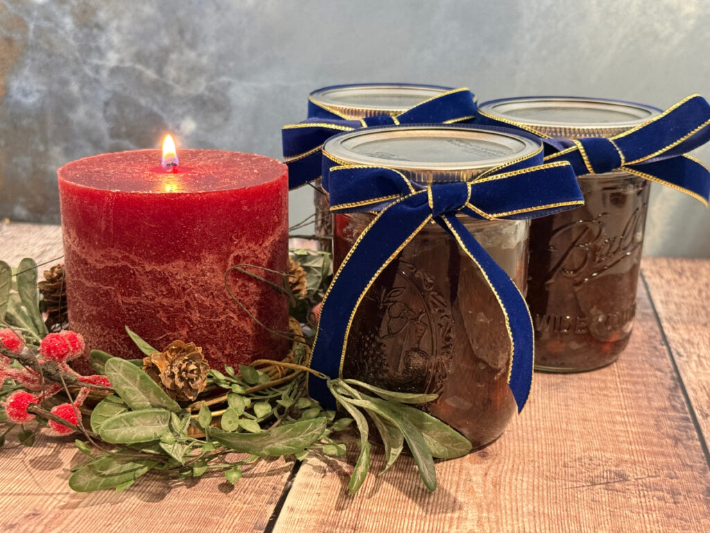 Three jars of suet-free mincemeat tied with blue and gold ribbon sitting beside a lit red candle encircled by Christmas foliage.