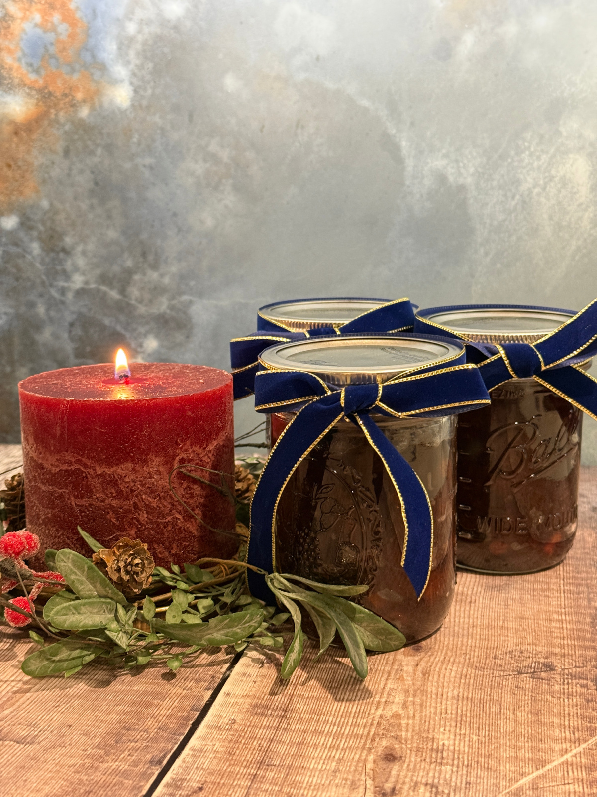 Three jars of suet-free mincemeat tied with blue and gold ribbon sitting beside a lit red candle surrounded by Christmas foliage