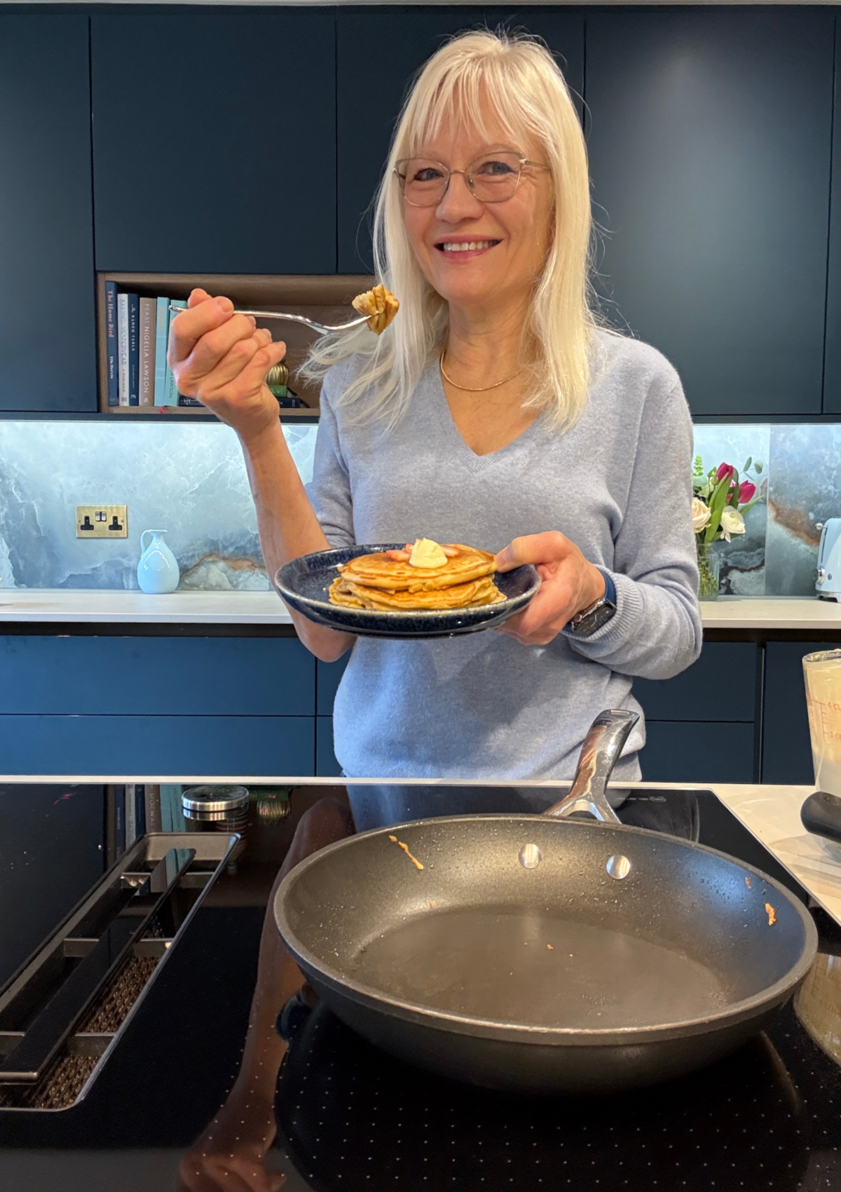April Harris standing by the stove top in her kitchen taking a bite from a stack of pancakes