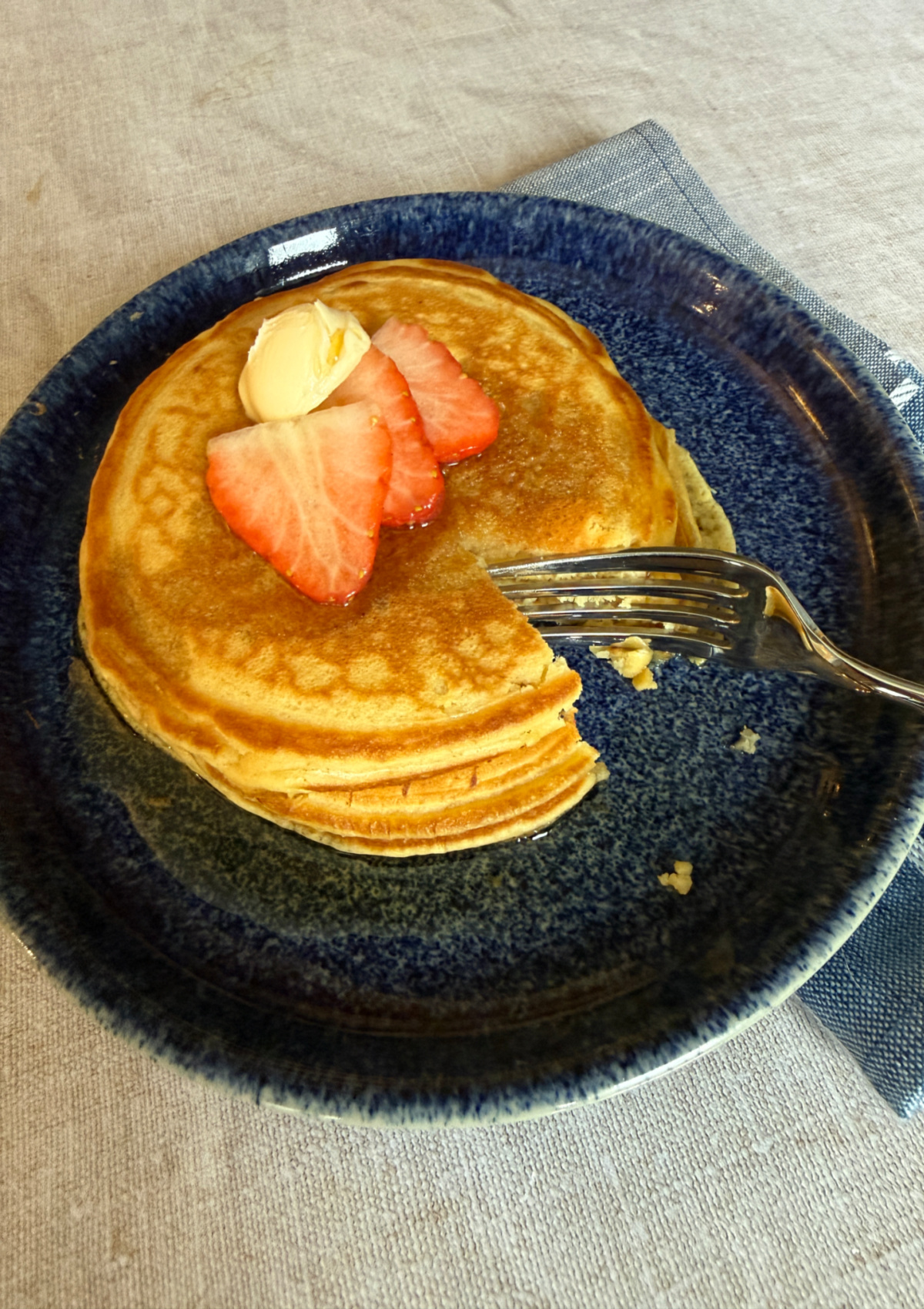 A stack of 3 dairy-free pancakes on a blue plate, topped with dairy-free spread, sliced strawberries and maple syrup. A bit is missing and there is a fork on the plate.