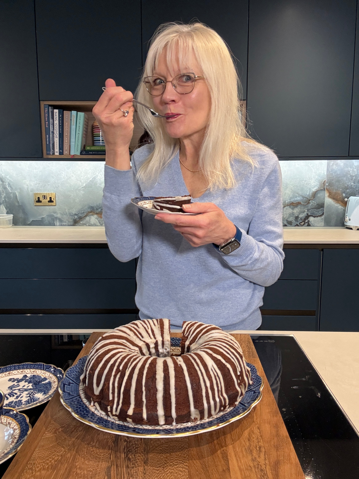 April Harris wearing a blue sweater, holding a plate with a piece of dairy-free chocolate cake on it. She is taking a bite of the slice from a fork. The whole cake minus the slice is in front of her on a Blue Willow china plate