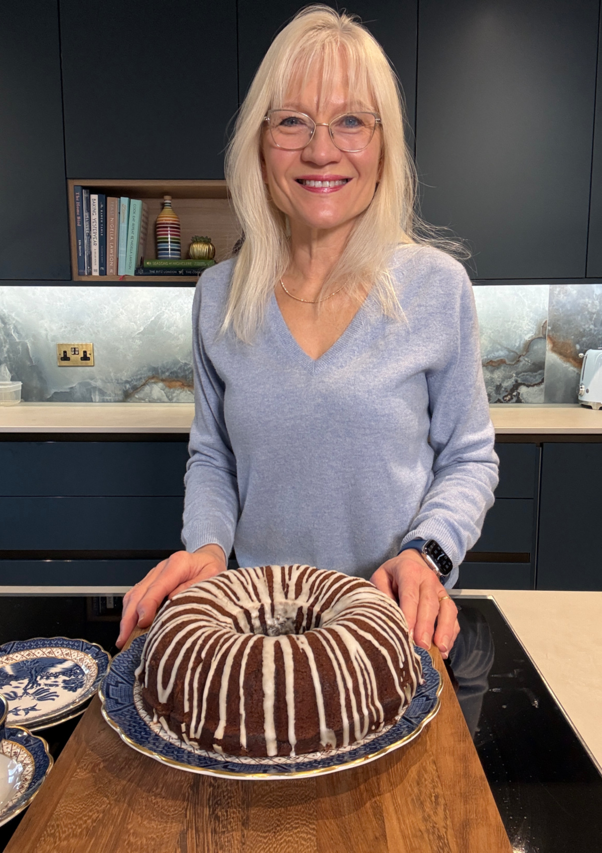 April Harris standing in her kitchen with a glazed dairy-free chocolate cake in front of her.