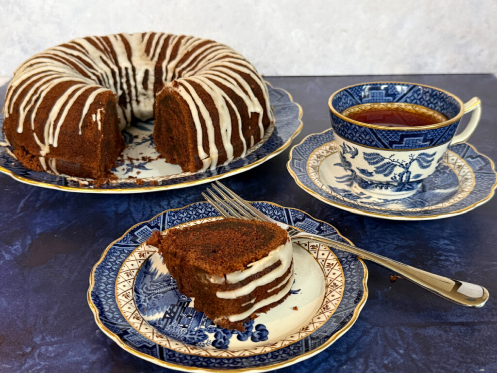 A dairy-free chocolate cake with a slice removed sitting on a Blue Willow China Plate with the cake slice on a plate in front and a cup of tea on the side