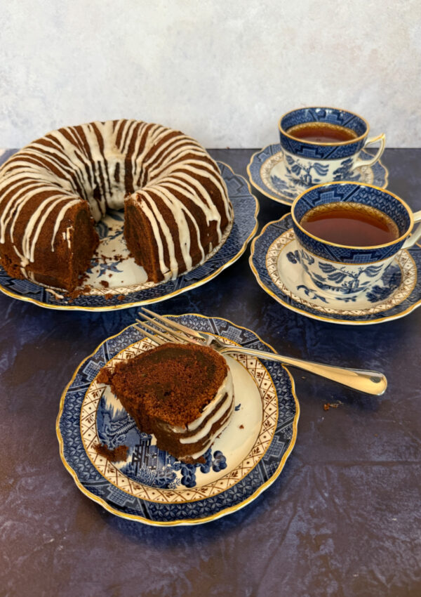 A dairy-free chocolate cake with a slice removed sitting on a Blue Willow China Plate with the cake slice on a plate in front and a cup of tea on the side