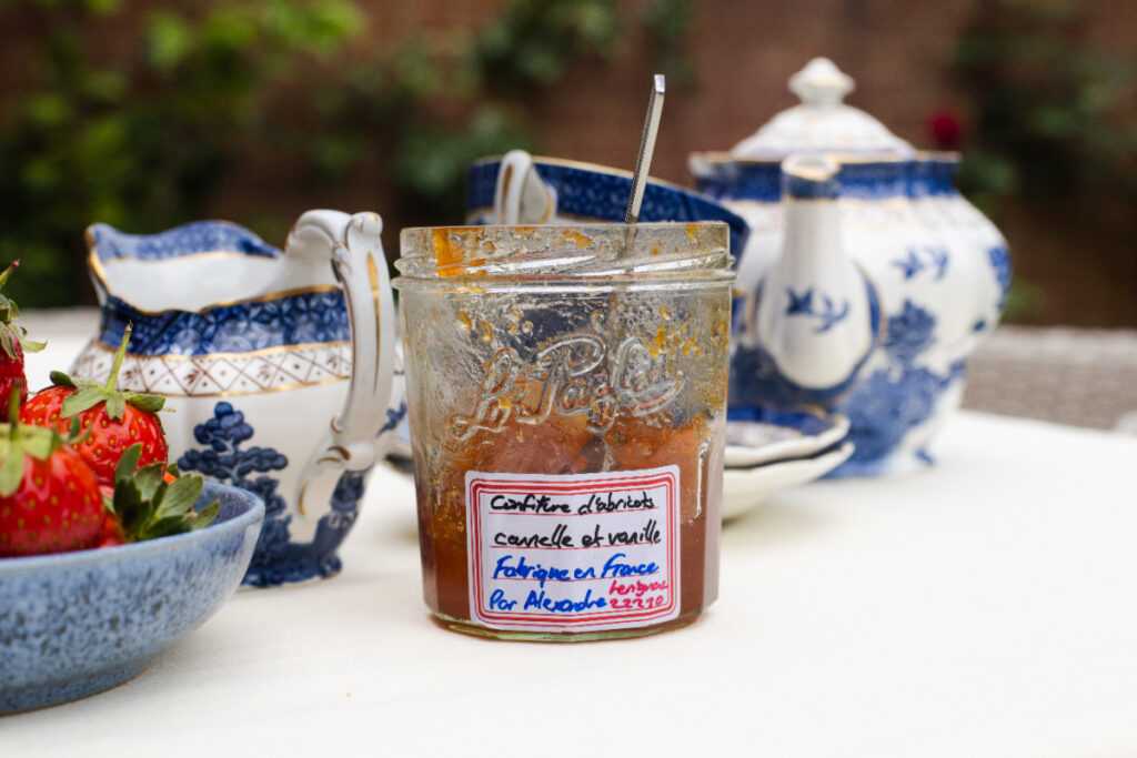 Homemade apricot jam with a handwritten label in front of Blue Willow china and beside a bowl of strawberries.
